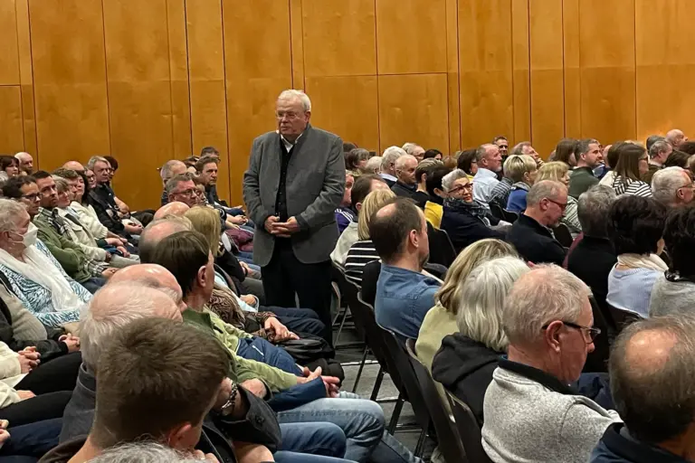 Ein älterer Mann steht inmitten von vielen sitzenden Personen