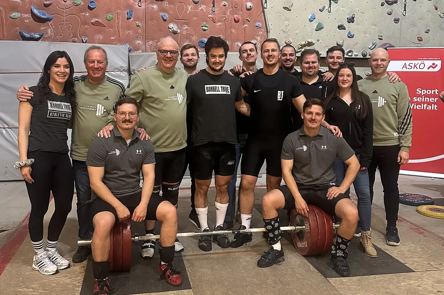 Eine Gruppe von Sportlerinnen und Sportlern posiert in einer Kletterhalle vor einer Wand mit Griffen. Zwei Männer sitzen vorne neben einer beladenen Langhantel, während die übrigen Teammitglieder dahinter stehen und in die Kamera lächeln.