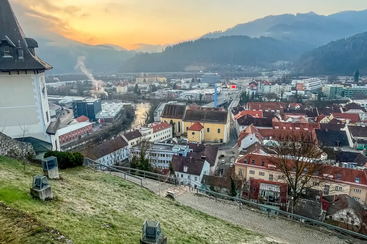 Blick über Gebäude und Straßen einer Stadt, am linken Bildrand ein Uhrturm, in der Bildmitte ein turmloser Kirchenbau mit gelber Fassade.