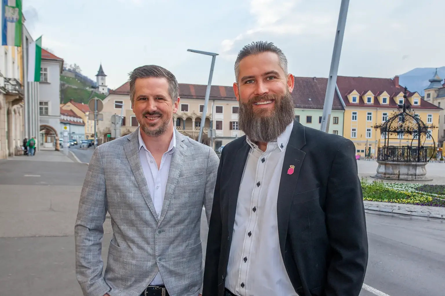 Zwei Männer in Hemd und Sakko, im Hintergrund die Brucker Altstadt mit Schloßberg, Kornmesserhaus und Eisernem Brunnen