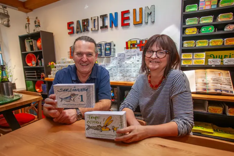Ein Mann und eine Frau präsentieren lächelnd Dosenfischverpackungen, im Hintergrund Regale mit weiteren Konserven