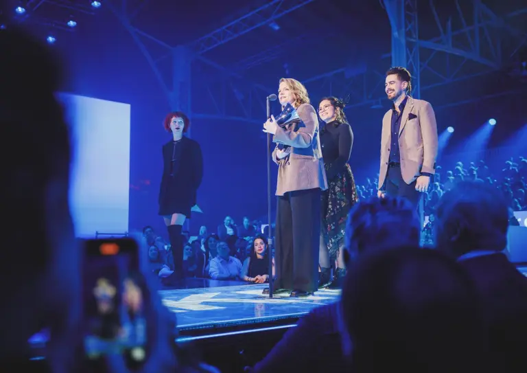 Drei Frauen und ein Mann auf einer Bühne. Eine der Frauen steht im Vordergrund an einem Mikrofon und hält eine Trophäe in Form eines Schalltrichters in Händen.
