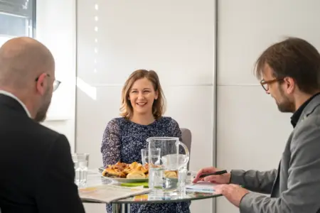 Eine Frau und zwei Männer unterhalten sich an einem runden Glastisch sitzend