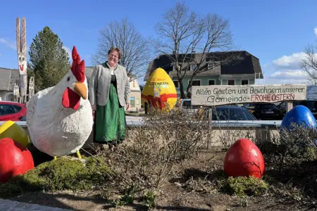 Eine Frau in Tracht steht lächelnd neben einer großen, bunt gestalteten Osterdekoration im Freien. Neben ihr sind ein übergroßes Huhn, bemalte Ostereier und ein dekorierter Platz mit Sträuchern zu sehen. Im Hintergrund steht ein Schild mit der Aufschrift „Allen Gästen und Bewohnern unserer Gemeinde – Frohe Ostern“, dahinter Häuser und parkende Autos unter blauem Himmel.
