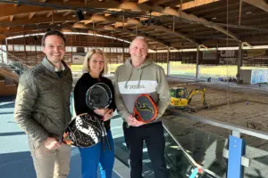 Zwei Männer und eine Frau mit Padelschlägern in einer großen Halle mit einem Bagger im Hintergrund