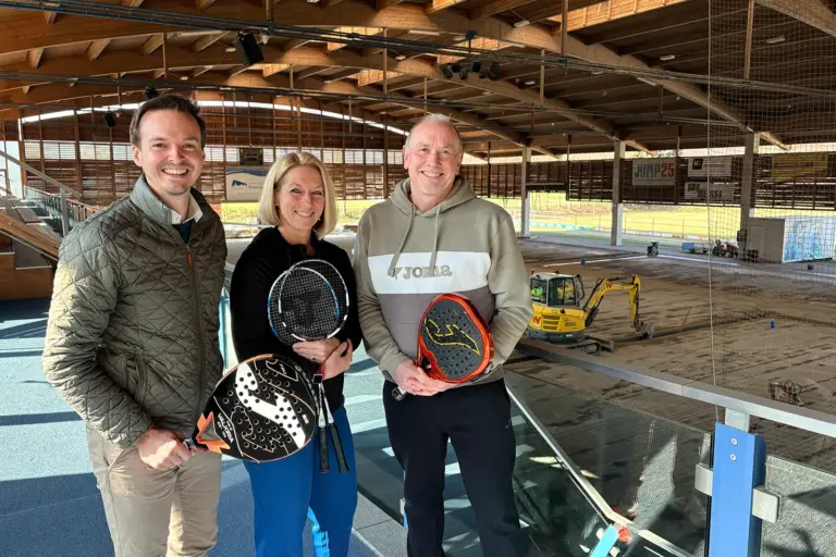 Bau der Ballsporthalle in Frohnleiten läuft Zwei Männer und eine Frau mit Padelschlägern in einer großen Halle mit einem Bagger im Hintergrund