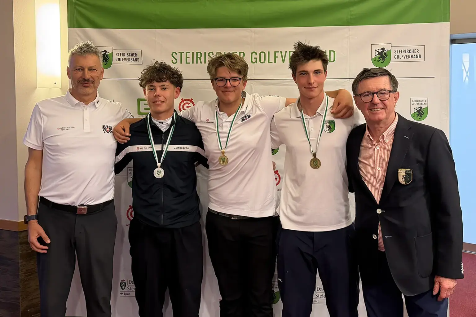 Fünf Männer stehen nebeneinander, die mittleren drei haben je eine Medaille um.