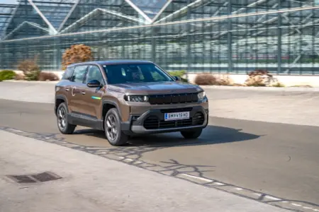 Ein braunes Auto, Karosserieform SUV, fährt von links nach rechts, im Hintergrund die Glasfassade eines großes Gewächshauses