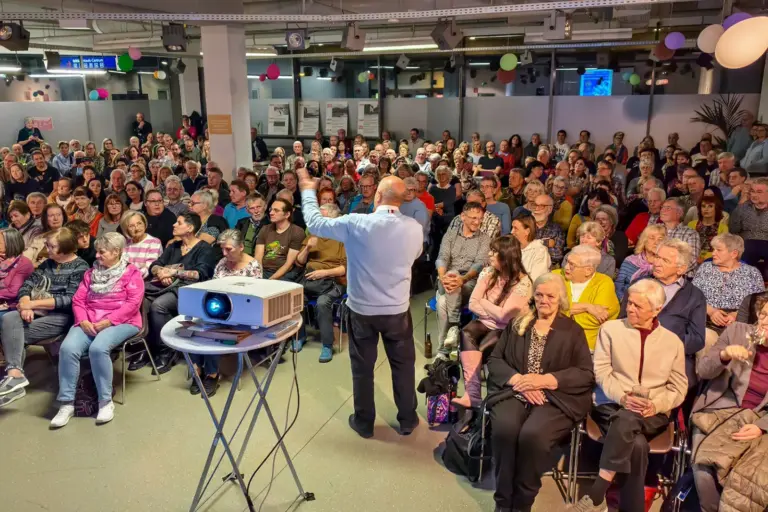 Ein Mann mit Mikrofon neben einem Projektor vor vielen ZuschauerInnen