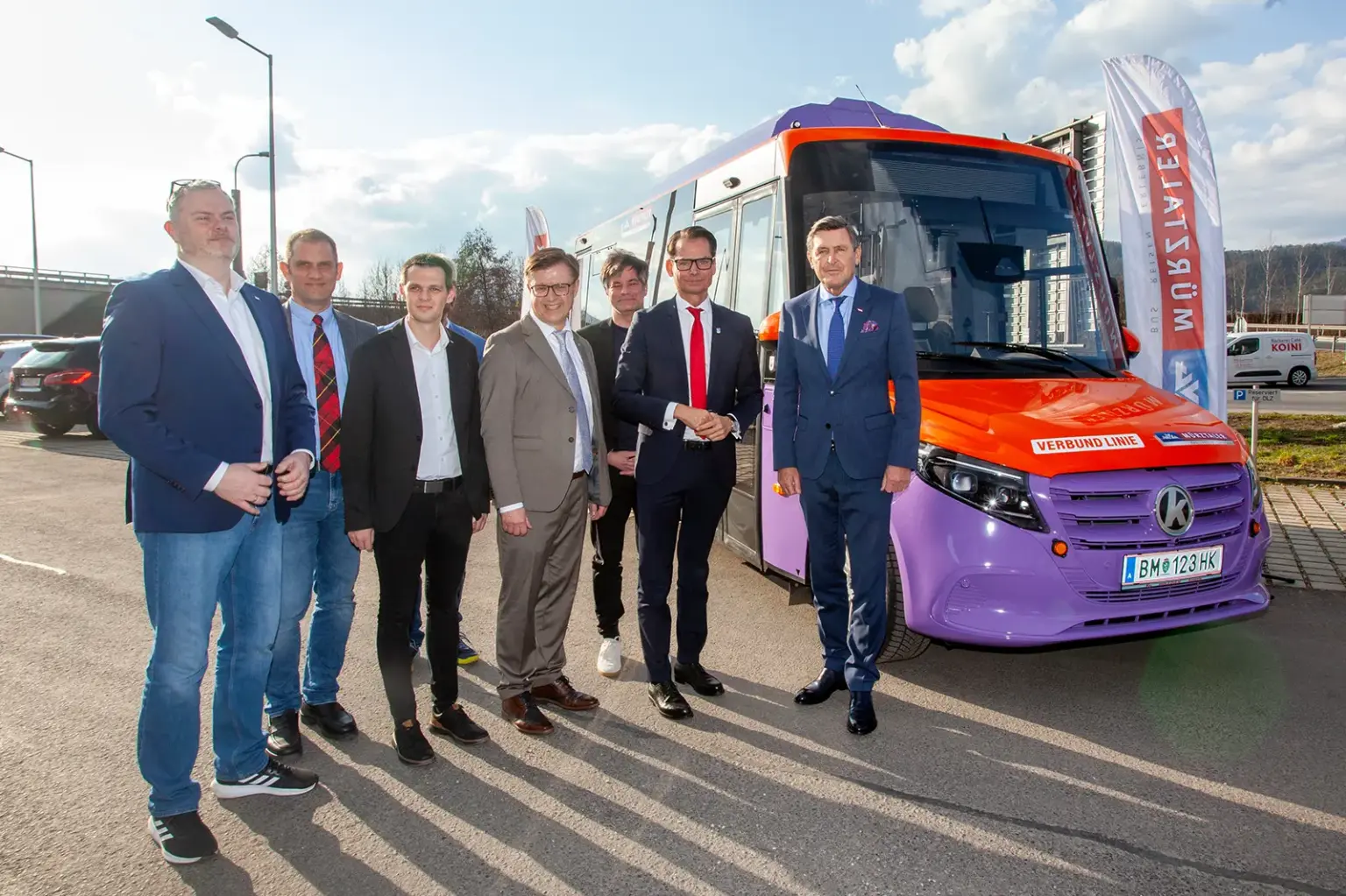 Sieben formell gekleidete Männer stehen neben einem in den Farben Lila und Orange lackierten Bus.