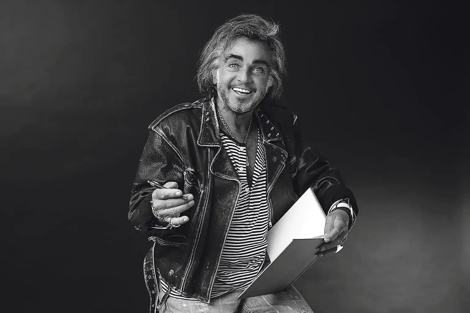 Ein Mann mit zirka schulterlangen Haaren und Fünftagebart in Lederjacke mit Skizzenblock und Stift in Händen