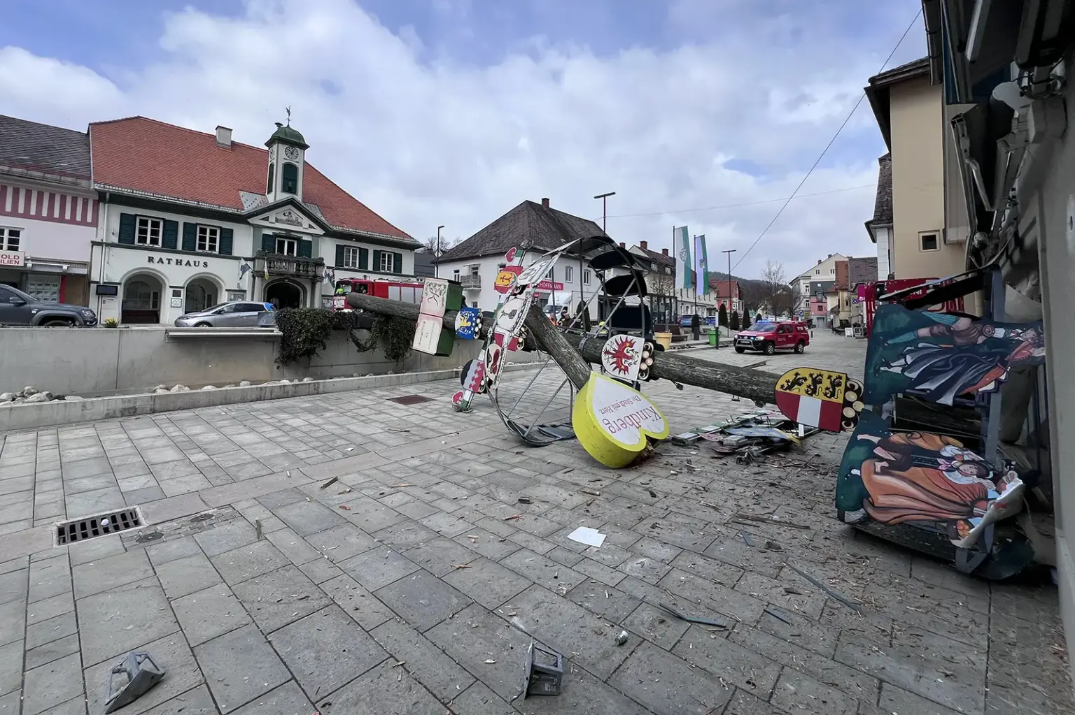 Ein umgestürzter, geschmückter Baum liegt auf einem Platz vor dem Rathaus, daneben verstreute Teile und Trümmer. Die dekorativen Tafeln am Stamm sind beschädigt, im Hintergrund sind Gebäude, Autos und Fahnen entlang der Straße zu sehen.