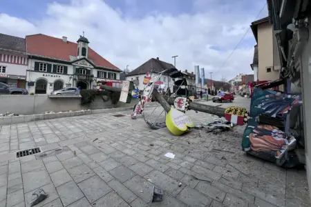 Ein umgestürzter, geschmückter Baum liegt auf einem Platz vor dem Rathaus, daneben verstreute Teile und Trümmer. Die dekorativen Tafeln am Stamm sind beschädigt, im Hintergrund sind Gebäude, Autos und Fahnen entlang der Straße zu sehen.