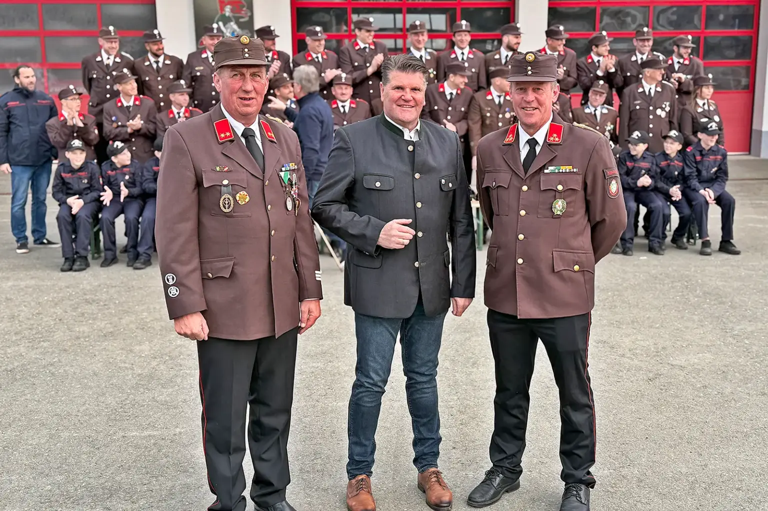 Zwei Männer in Uniform und einer in Trachtenjacke vor einer Feuerwehrmannschaft