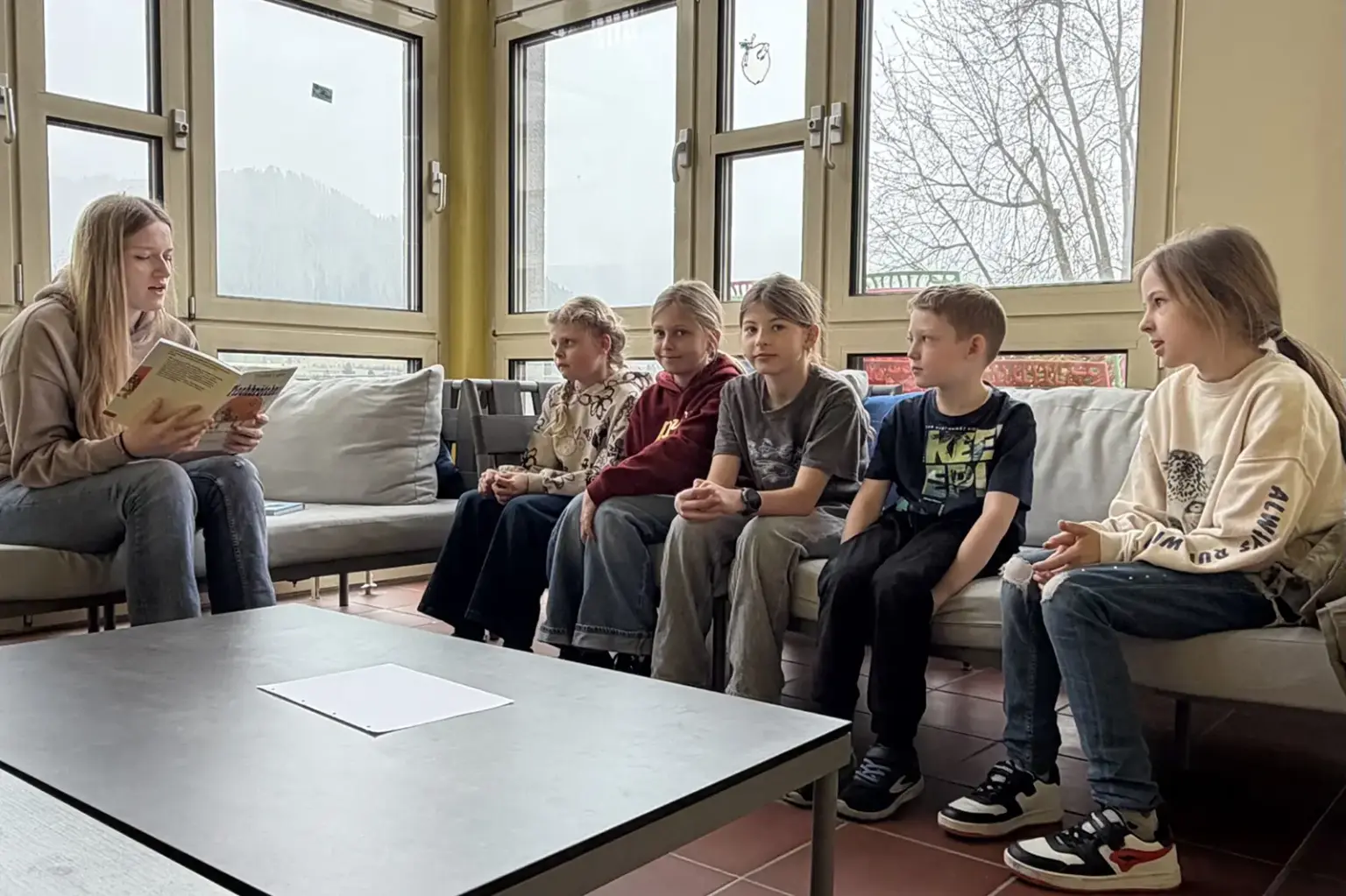 Eine Jugendlche sitzt mit fünf Kindern auf einer Couch und liest ihnen vor