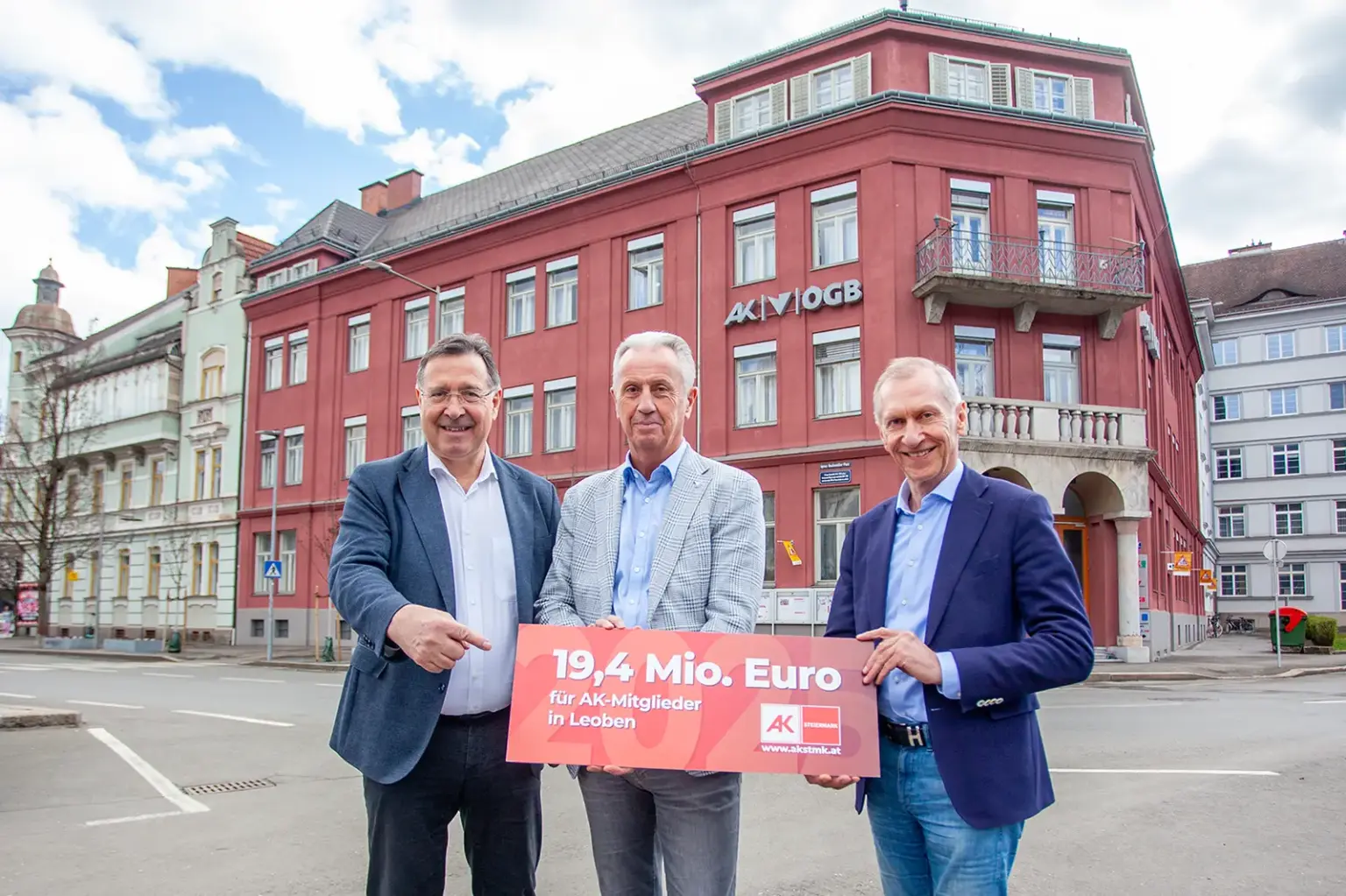 Drei Männer in formeller Kleidung stehen vor einem Haus mit roter Fassade, in den Händen ein Schild mit der Aufschrift "19,4 Mio Euro"