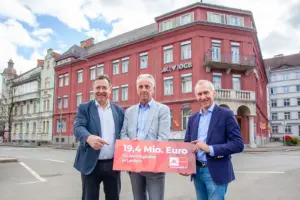 Drei Männer in formeller Kleidung stehen vor einem Haus mit roter Fassade, in den Händen ein Schild mit der Aufschrift "19,4 Mio Euro"