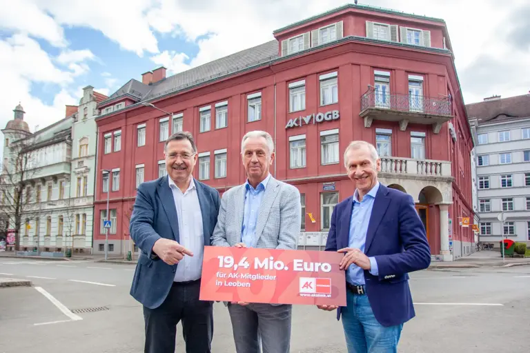 Drei Männer in formeller Kleidung stehen vor einem Haus mit roter Fassade, in den Händen ein Schild mit der Aufschrift "19,4 Mio Euro"