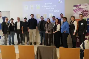 Ein Gruppe von Menschen stehen nebeneinander in einem Raum. Hinter ihnen an der Wand ist eine Präsentation zu sehen. Vor ihnen stehen Sesseln.