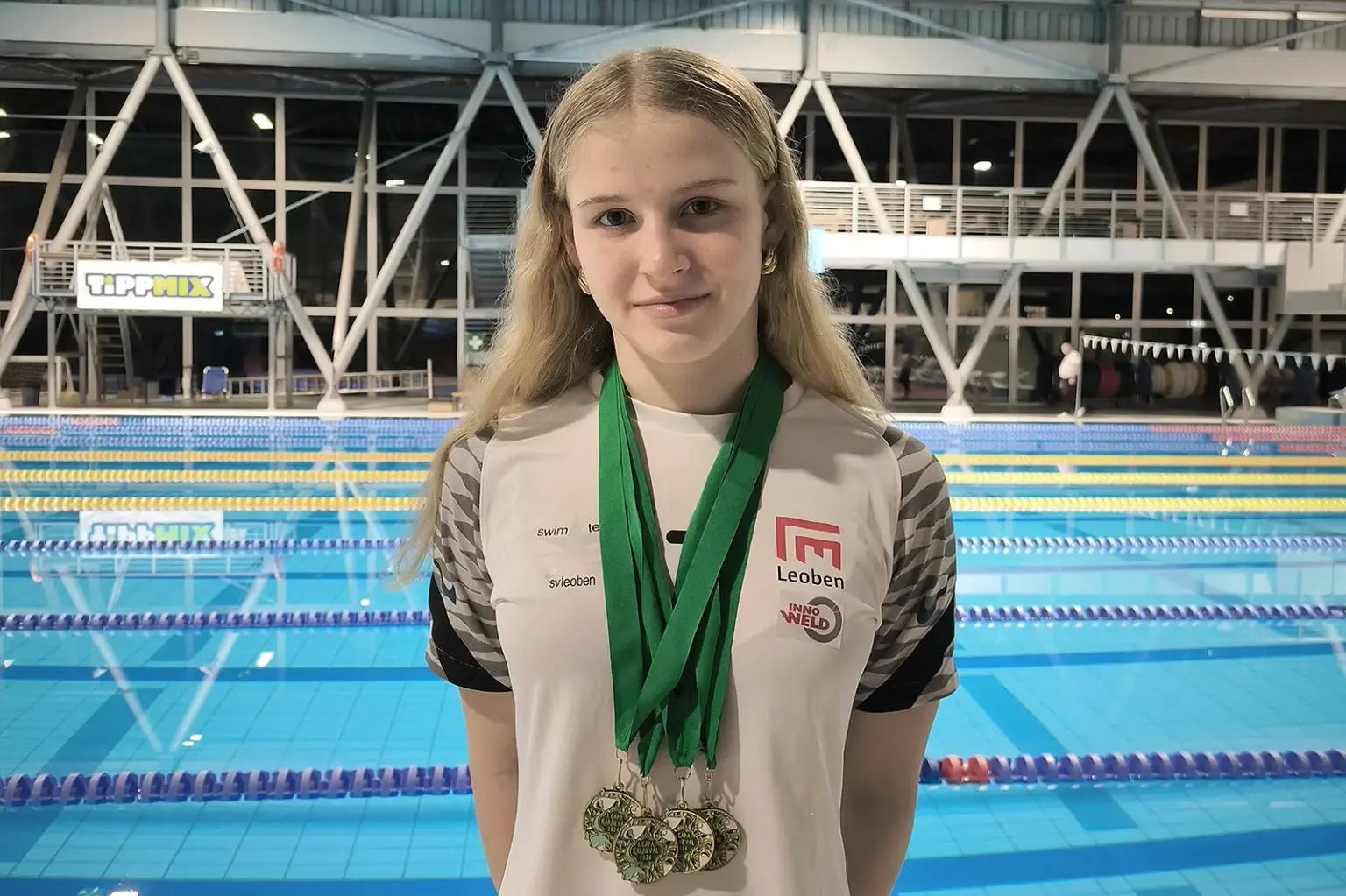 Lilli Paier knackte das Limit für die Junioren-EM Eine junge Dame mit vier Medaillen um den Hals, hinter ihr ist ein Hallen-Schwimmbad erkennbar.