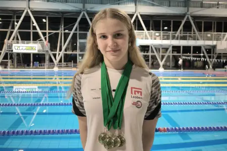 Eine junge Dame mit vier Medaillen um den Hals, hinter ihr ist ein Hallen-Schwimmbad erkennbar.