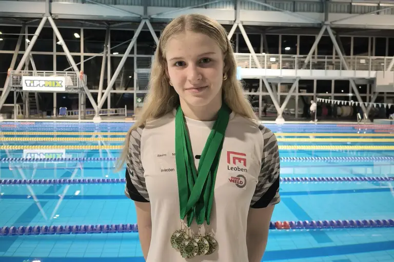 Eine junge Dame mit vier Medaillen um den Hals, hinter ihr ist ein Hallen-Schwimmbad erkennbar.