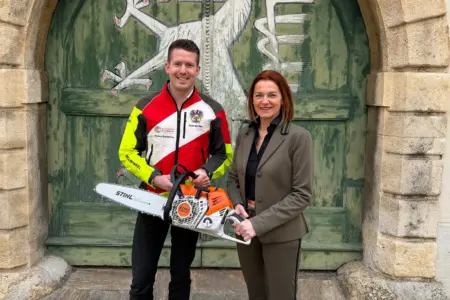 Ein Mann und eine Frau mit einer Kettensäge vor einem Holztor mit dem steirischen Panther