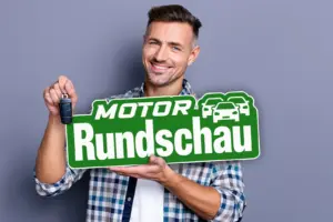Ein junger Mann hält einen Autoschlüssel und eine Tafel mit dem Schriftzug "Motor Rundschau" in den Händen