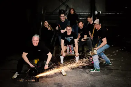 Eine Gruppe von acht Personen posiert in einer Werkstatt mit Werkzeugen in den Händen. Im Vordergrund erzeugt ein Mann mit einer Flex Funken, während die anderen mit Hämmern, Schraubenziehern und anderen Geräten in die Kamera blicken. Die Szene wirkt inszeniert und dynamisch, mit dunklem Hintergrund und dramatischer Beleuchtung.