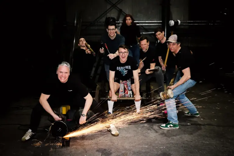Eine Gruppe von acht Personen posiert in einer Werkstatt mit Werkzeugen in den Händen. Im Vordergrund erzeugt ein Mann mit einer Flex Funken, während die anderen mit Hämmern, Schraubenziehern und anderen Geräten in die Kamera blicken. Die Szene wirkt inszeniert und dynamisch, mit dunklem Hintergrund und dramatischer Beleuchtung.