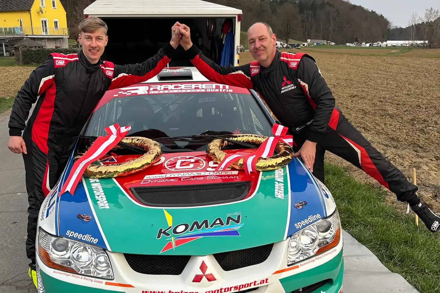 Zwei Männer in Rennanzügen stehen vor einem Rallyeauto und schlagen die Hände über dem Dach ein. Auf der Motorhaube liegen zwei Siegerkränze mit rot-weiß-roten Bändern. Das Auto ist mit Sponsorenlogos beklebt, im Hintergrund sind ein Haus, Felder und ein offenes Garagentor zu sehen.