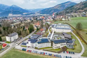 Luftaufnahme einer ländlichen Gemeinde in bergiger Gegend, im Vordergrund ein Komplex aus größeren Gebäuden