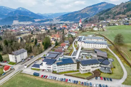 Luftaufnahme einer ländlichen Gemeinde in bergiger Gegend, im Vordergrund ein Komplex aus größeren Gebäuden