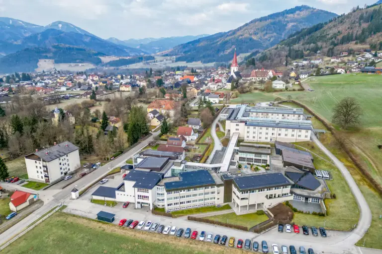 Luftaufnahme einer ländlichen Gemeinde in bergiger Gegend, im Vordergrund ein Komplex aus größeren Gebäuden