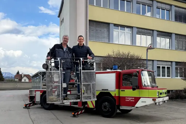 Zwei Männer im Hebekorb eines alten Feuerwehrfahrzeuges vor einem Haus
