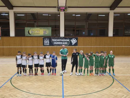 Kinder-Fußballteams stehen in einer Turnhalle nebeneinander aufgestellt, links in weißen und rechts in grünen Trikots. In der Mitte steht ein Trainer mit Ball. Im Hintergrund hängen Vereinsbanner an der Holzwand, darunter „Teichstadion Hurricans“.