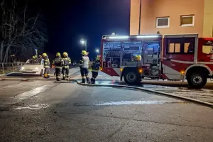 Ein Kleinwagen steht bei Nacht auf einer beleuchteten Straße, ringsum bzw. daneben acht Feuerwehrleute in Einsatzkleidung und ein Rüstfahrzeug, quer über die Straße liegt ein Löschschlauch.