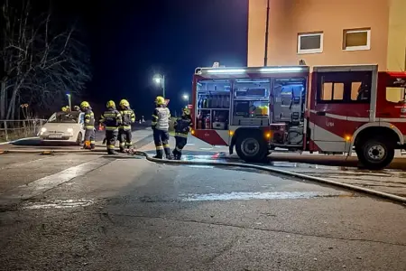 Ein Kleinwagen steht bei Nacht auf einer beleuchteten Straße, ringsum bzw. daneben acht Feuerwehrleute in Einsatzkleidung und ein Rüstfahrzeug, quer über die Straße liegt ein Löschschlauch.