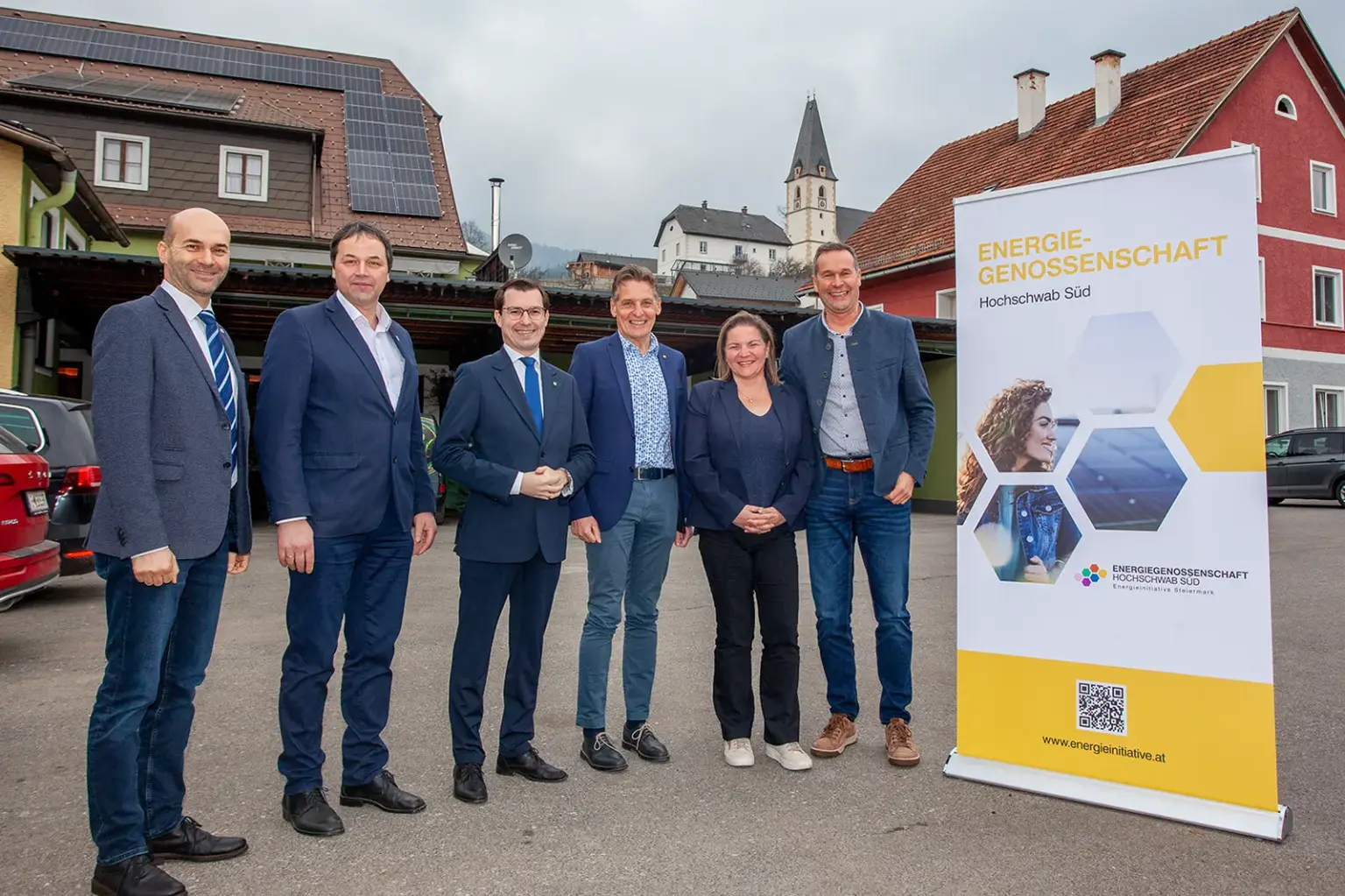 Fünf Männer und eine Frau stehen im Freien neben einem Werbeaufsteller mit der Aufschrift "Energiegenossenschaft Hochschwab Süd", im Hintergrund ein Dach mit Photovoltaikanlage und die Kirche von Turnau