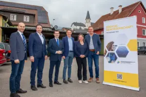 Fünf Männer und eine Frau stehen im Freien neben einem Werbeaufsteller mit der Aufschrift "Energiegenossenschaft Hochschwab Süd", im Hintergrund ein Dach mit Photovoltaikanlage und die Kirche von Turnau