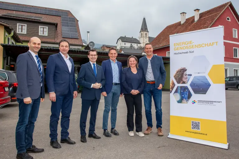 Fünf Männer und eine Frau stehen im Freien neben einem Werbeaufsteller mit der Aufschrift "Energiegenossenschaft Hochschwab Süd", im Hintergrund ein Dach mit Photovoltaikanlage und die Kirche von Turnau