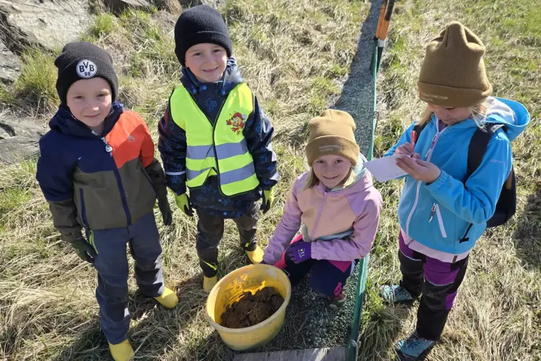 Vier Kinder stehen auf einer Wiese neben einem Eimer