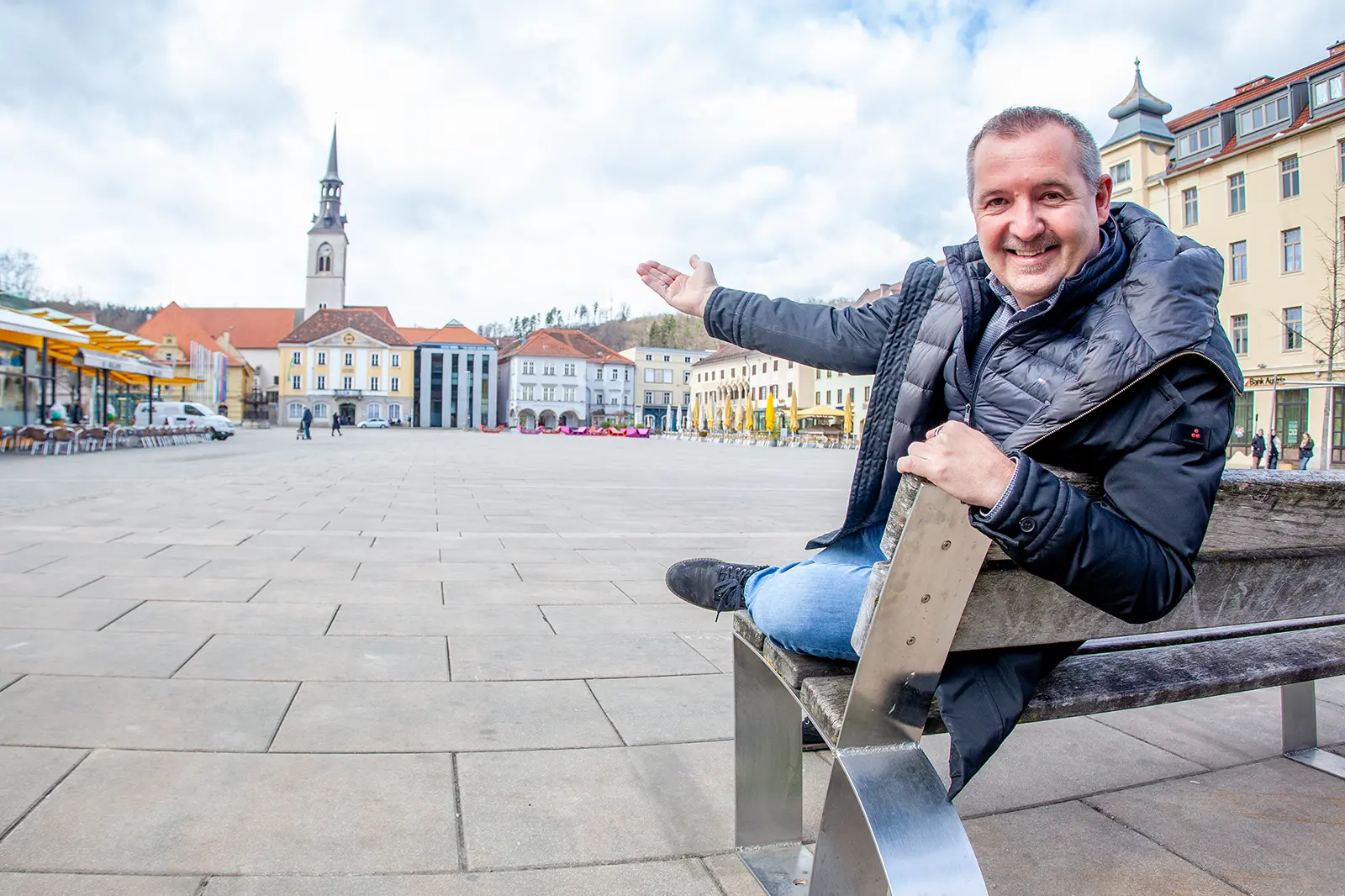 Ein Mann sitzt auf einer Bank und zeigt auf einen großen innerstädtischen Platz