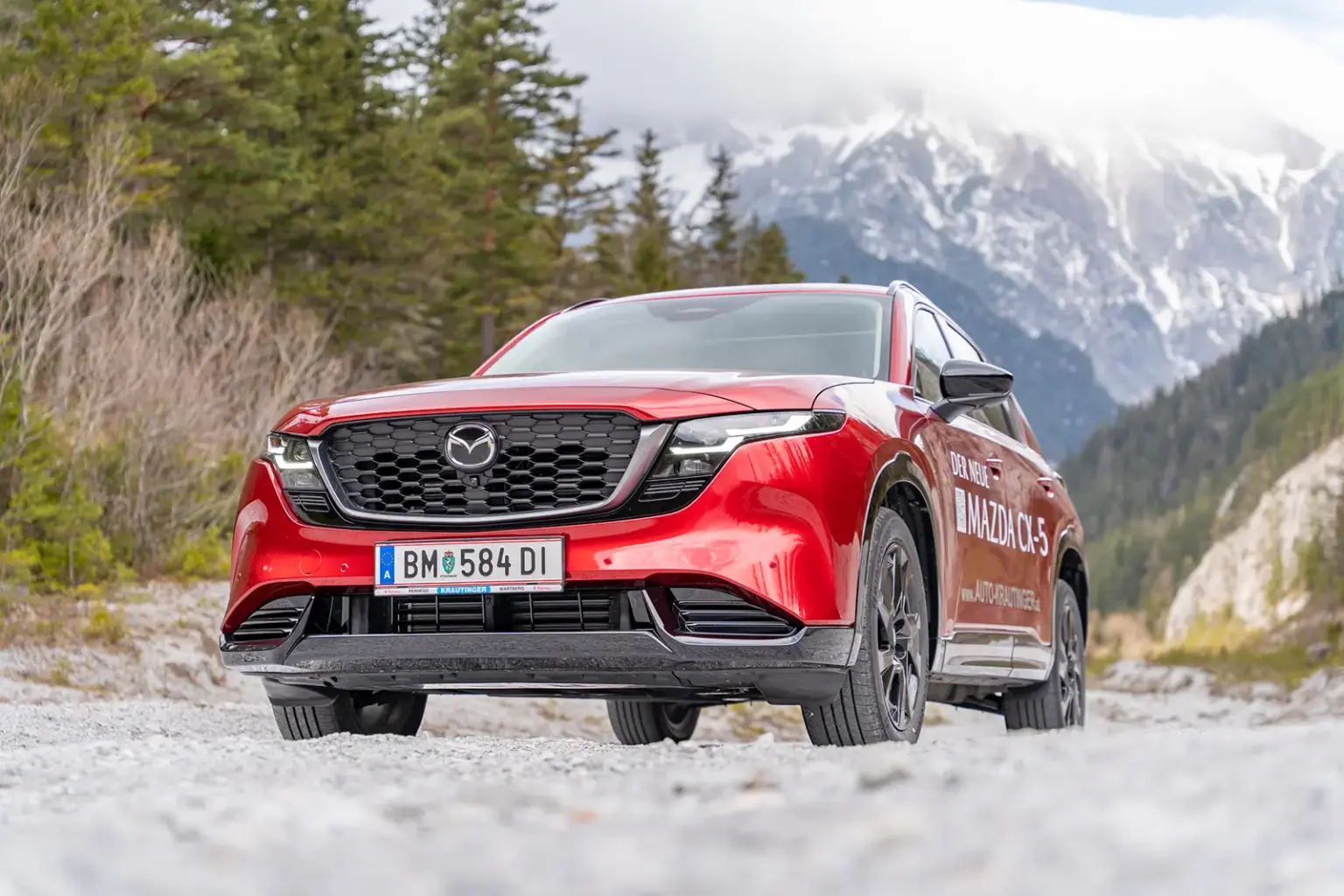 Ein roter Pkw steht auf einer Schotterfläche, im Hintergrund Wald und eine Felswand, nach oben hin in Wolken gehüllt