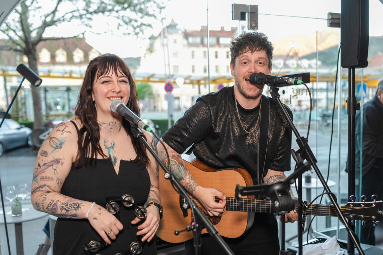 Eine Frau mit Percussioninstrument und ein Mann mit Gitarre singen in einem Lokal, im Hintergrund ein innerstädtischer Platz