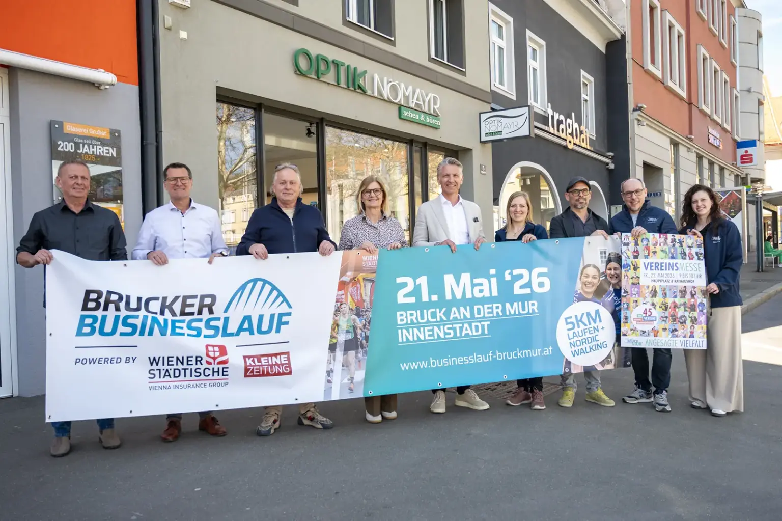 Eine Gruppe von Personen steht vor Geschäften in der Innenstadt und hält Banner für den „Brucker Businesslauf“. Auf den Plakaten sind Termin und Veranstaltungsdetails zu sehen, die Beteiligten präsentieren die Veranstaltung gemeinsam auf der Straße.