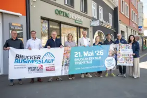 Eine Gruppe von Personen steht vor Geschäften in der Innenstadt und hält Banner für den „Brucker Businesslauf“. Auf den Plakaten sind Termin und Veranstaltungsdetails zu sehen, die Beteiligten präsentieren die Veranstaltung gemeinsam auf der Straße.