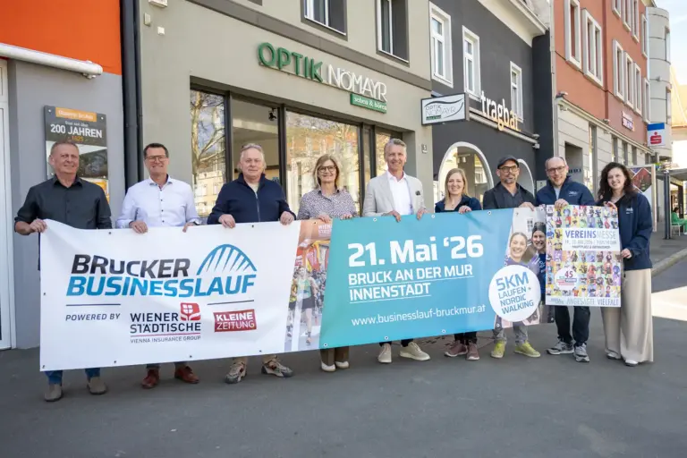 Eine Gruppe von Personen steht vor Geschäften in der Innenstadt und hält Banner für den „Brucker Businesslauf“. Auf den Plakaten sind Termin und Veranstaltungsdetails zu sehen, die Beteiligten präsentieren die Veranstaltung gemeinsam auf der Straße.