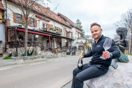 Ein Mann sitzt mit einer Flasche und einem Weinglas in Händen auf einem Stein neben der Statuette einer Nixe, im Hintergrund ein Gastgarten und ein Haus mit historischer Fassade