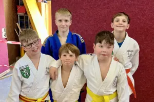 Fünf Kinder in Judoanzügen stehen eng beisammen und posieren lächelnd für ein Gruppenfoto in einer Turnhalle.